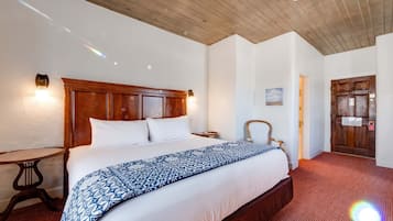 A hotel room with a wooden headboard, a large bed with a patterned bedspread, a small round table, and a door with a sign.
