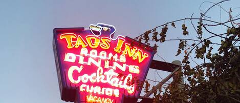 Letreiro de neon do 'Taus Inn' com os serviços 'Serviço de Quartos' e 'Cocktail', e um aviso de 'Vagas'.