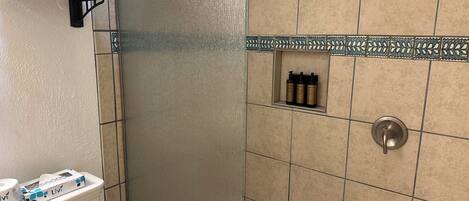 Un baño con baldosas color beige, una ducha, un inodoro y un estante con artículos de aseo.