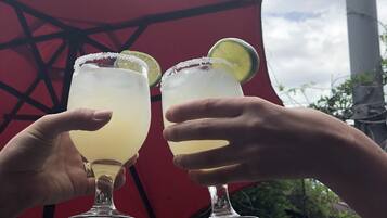 Two hands holding margaritas with lime slices, a sign reading "Tacos Inn", and a red umbrella in the background.