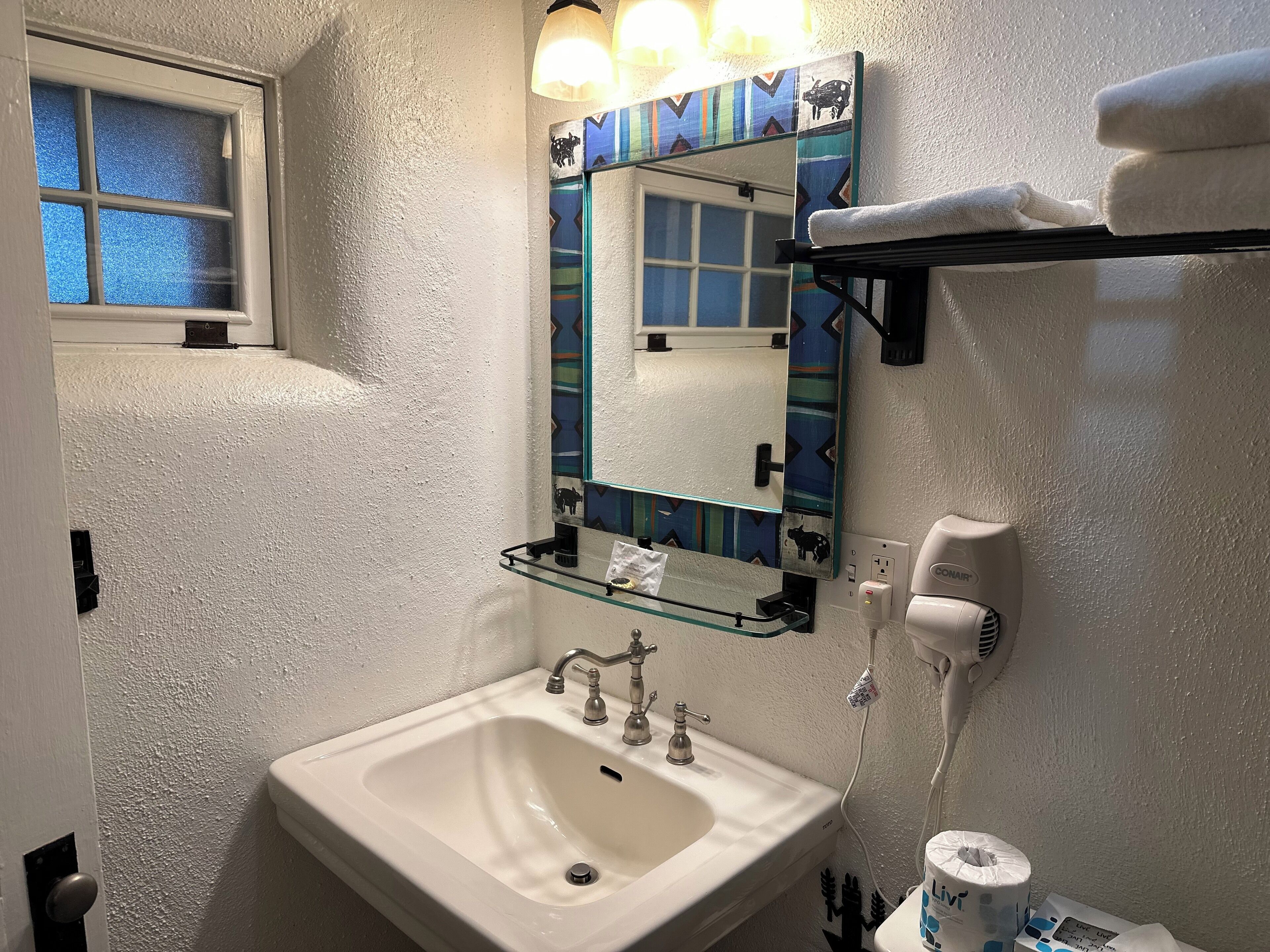 A bathroom with a white sink, a mirror with a decorative frame, a towel rack with a towel, and a hairdryer on the wall.