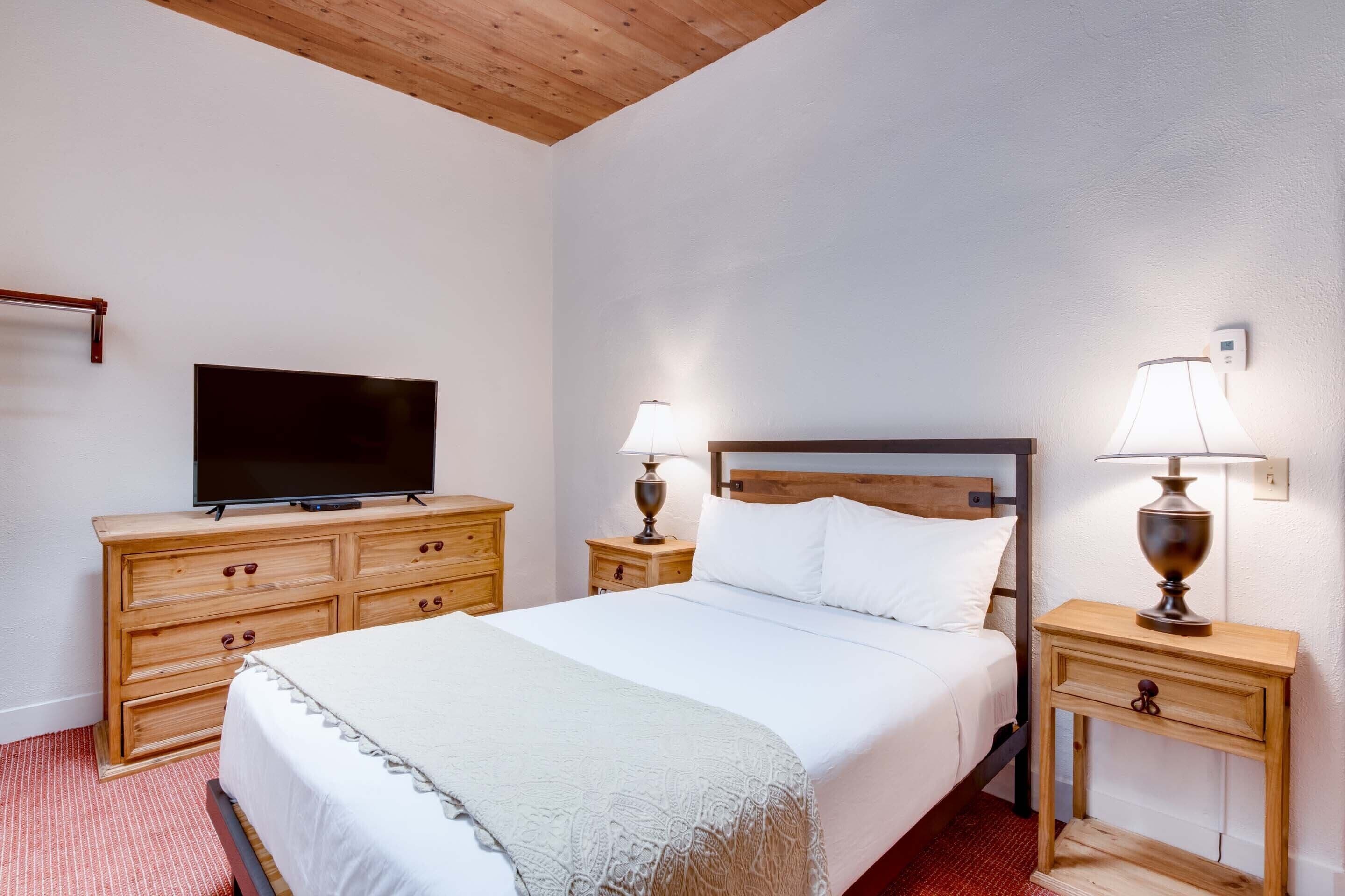 A hotel room with a bed, wooden dresser, TV, two bedside lamps, and a red carpet.