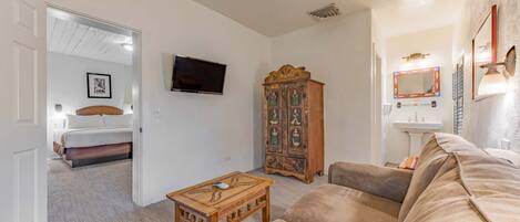 A living room with a beige sofa, a wooden coffee table, a television, a cabinet with artwork, and a doorway leading to a bedroom.