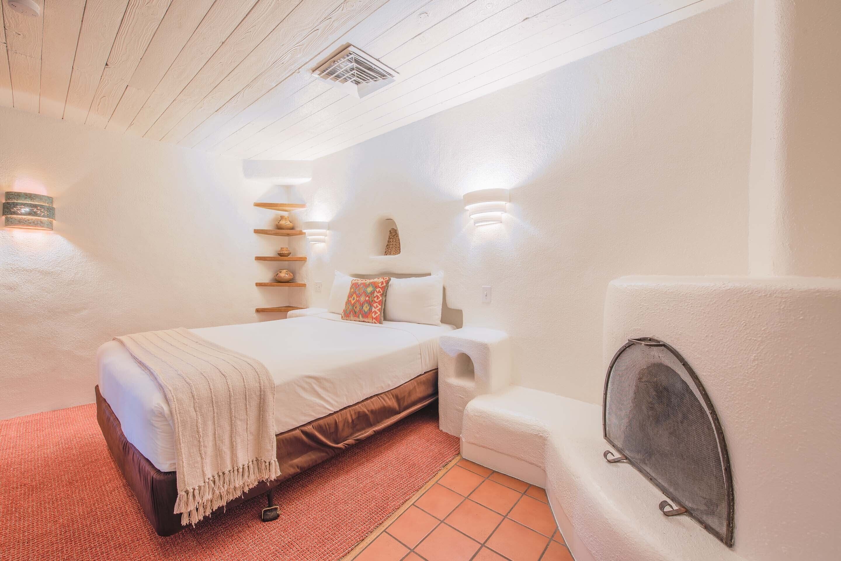 Un dormitorio con cama, una pequeña zona de estar, una estantería con adornos y una pantalla montada en la pared.