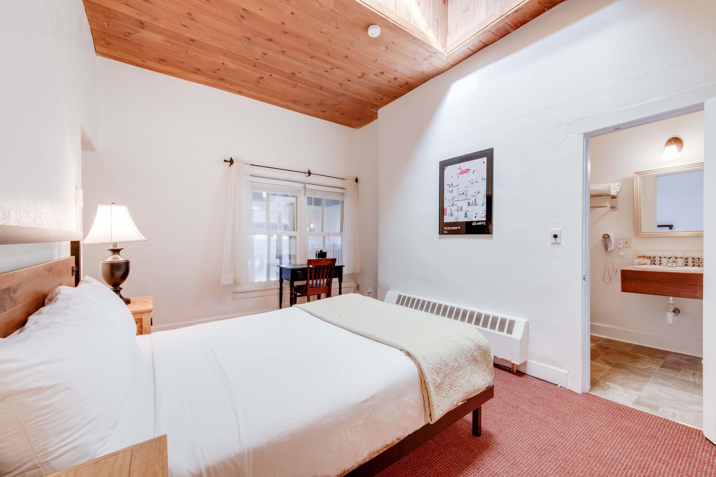 A bedroom with a wooden ceiling, a bed, a desk, a chair, a window, a radiator, and a bathroom with a mirror and sink.
