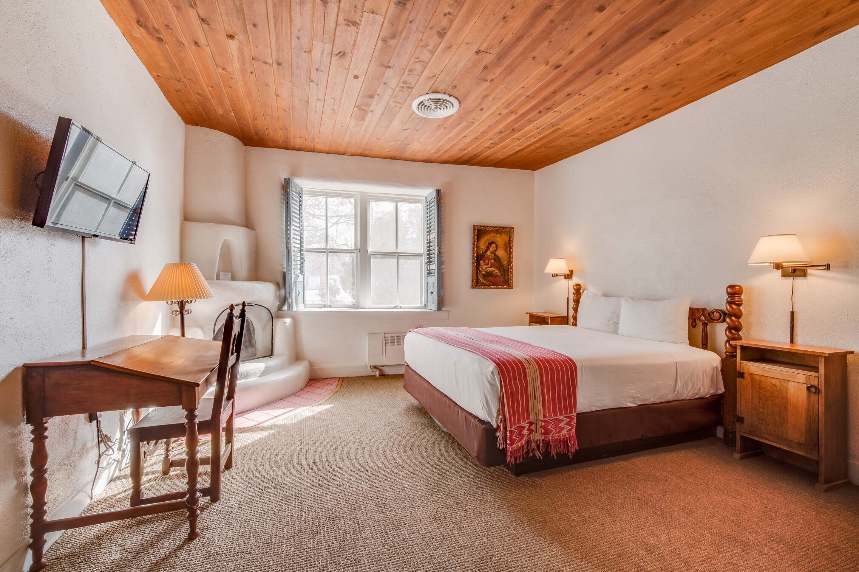 Un dormitorio con techo de madera, una cama con manta roja, un escritorio con silla, un televisor montado en la pared y una ventana con contraventanas.