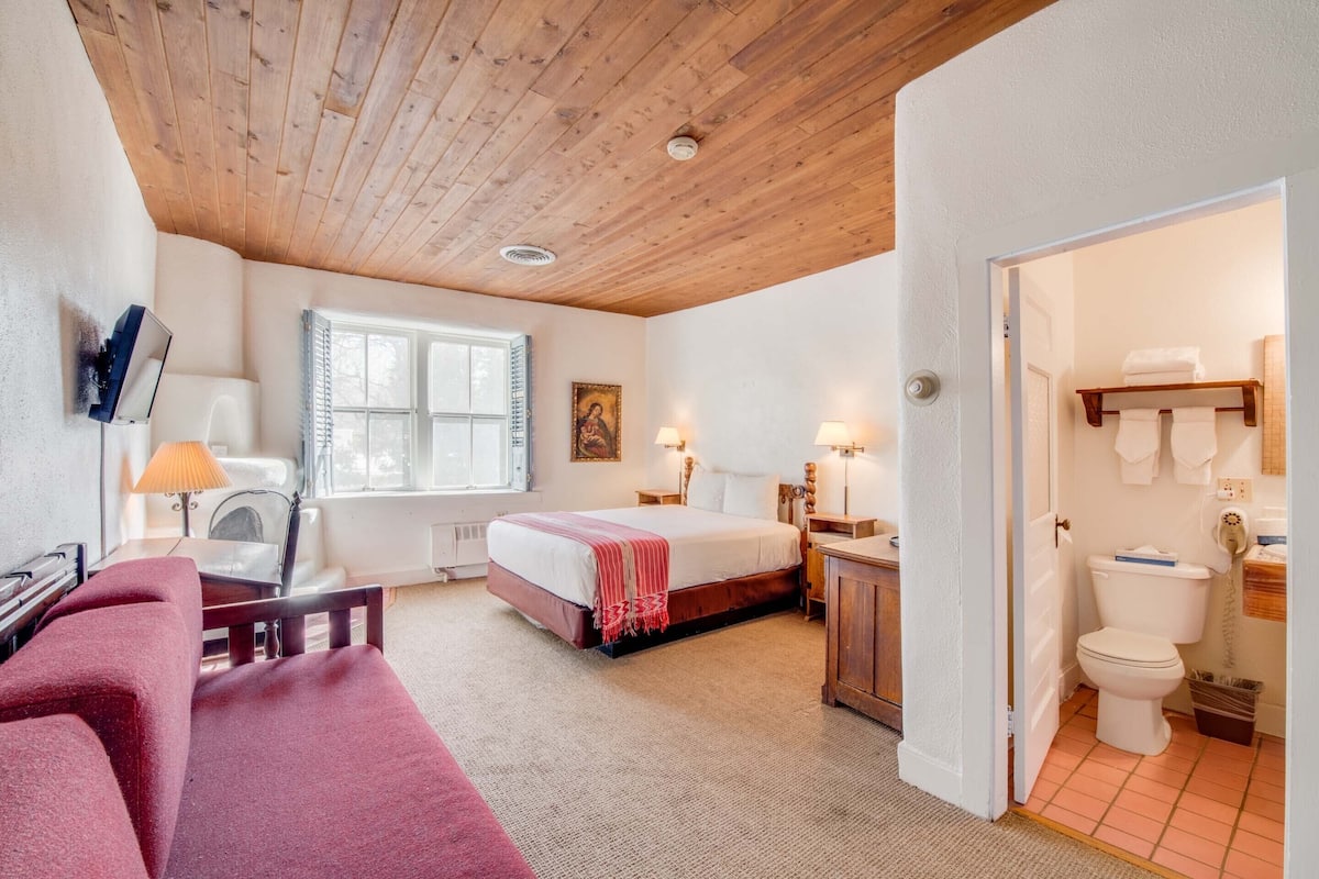 A cozy bedroom with a wooden ceiling, a bed, a desk with a lamp, a chair, and a window.