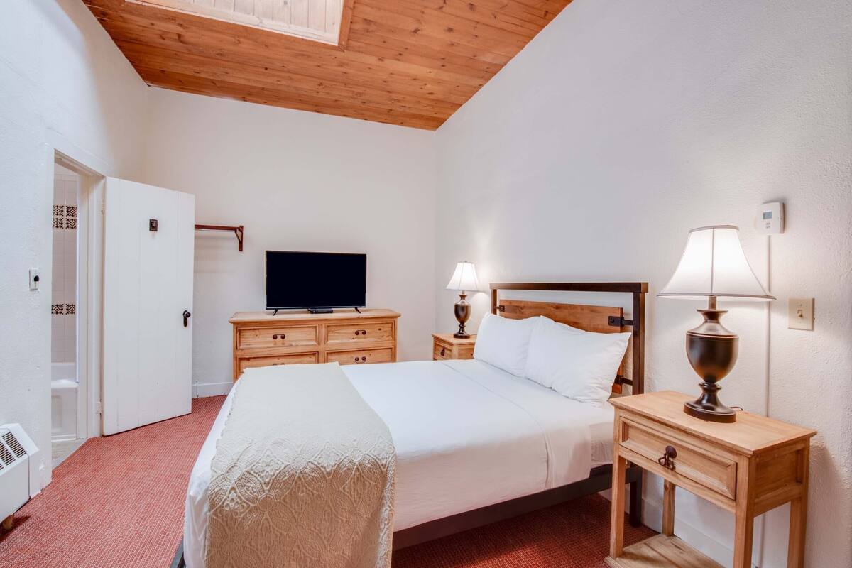 A hotel room with a wooden ceiling, a bed with white linens, two bedside tables with lamps, a TV on a dresser, and a door leading to a bathroom.