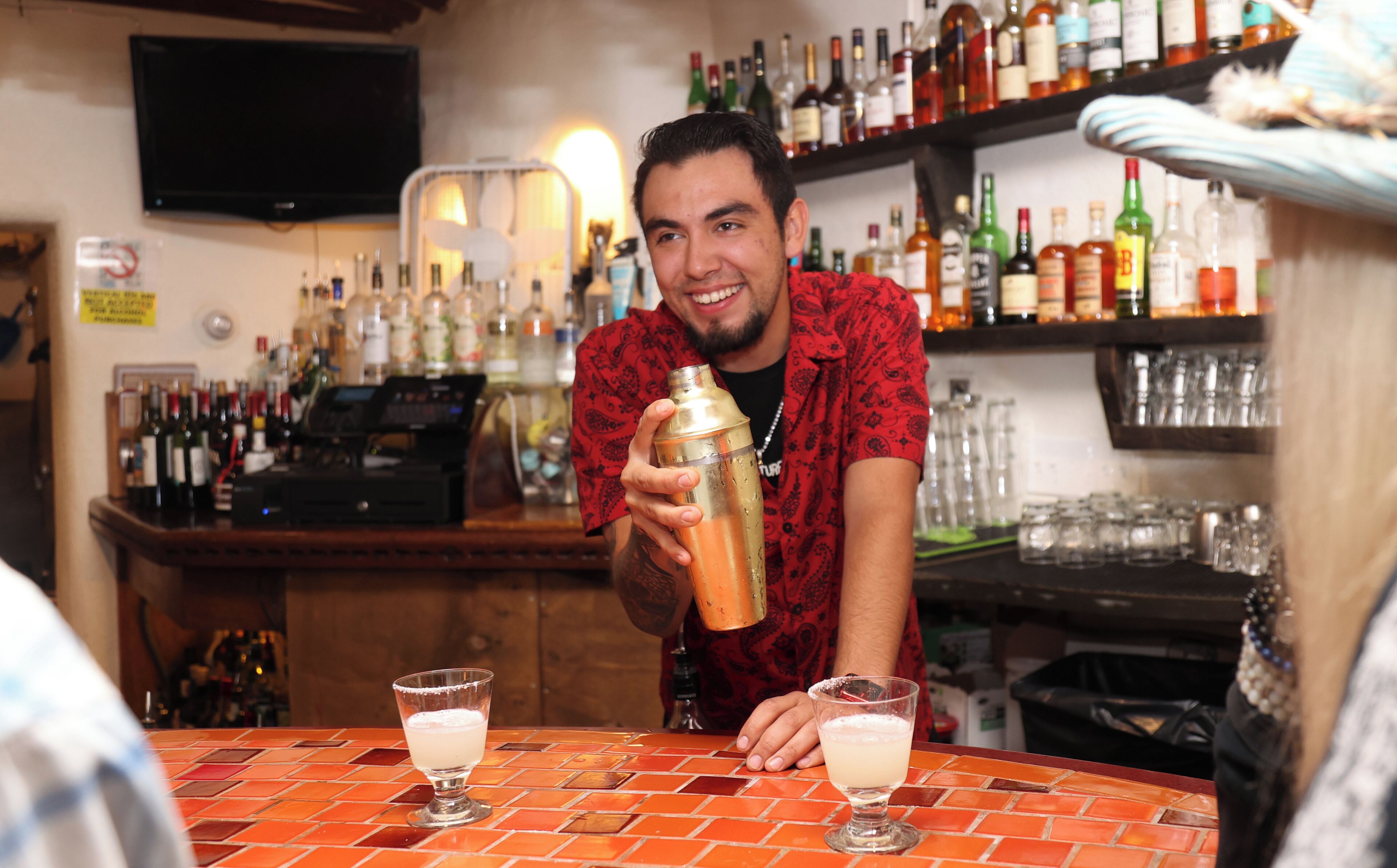 A bar with a bartender preparing drinks. There are multiple bottles and glasses on shelves behind the bartender.