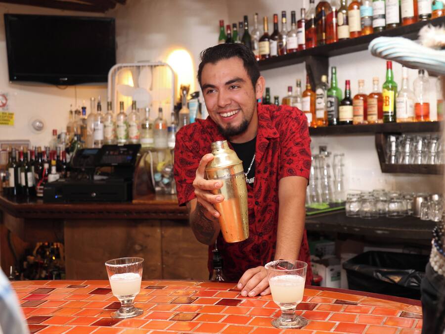 A bar with a bartender preparing drinks. There are multiple bottles and glasses on shelves behind the bartender.