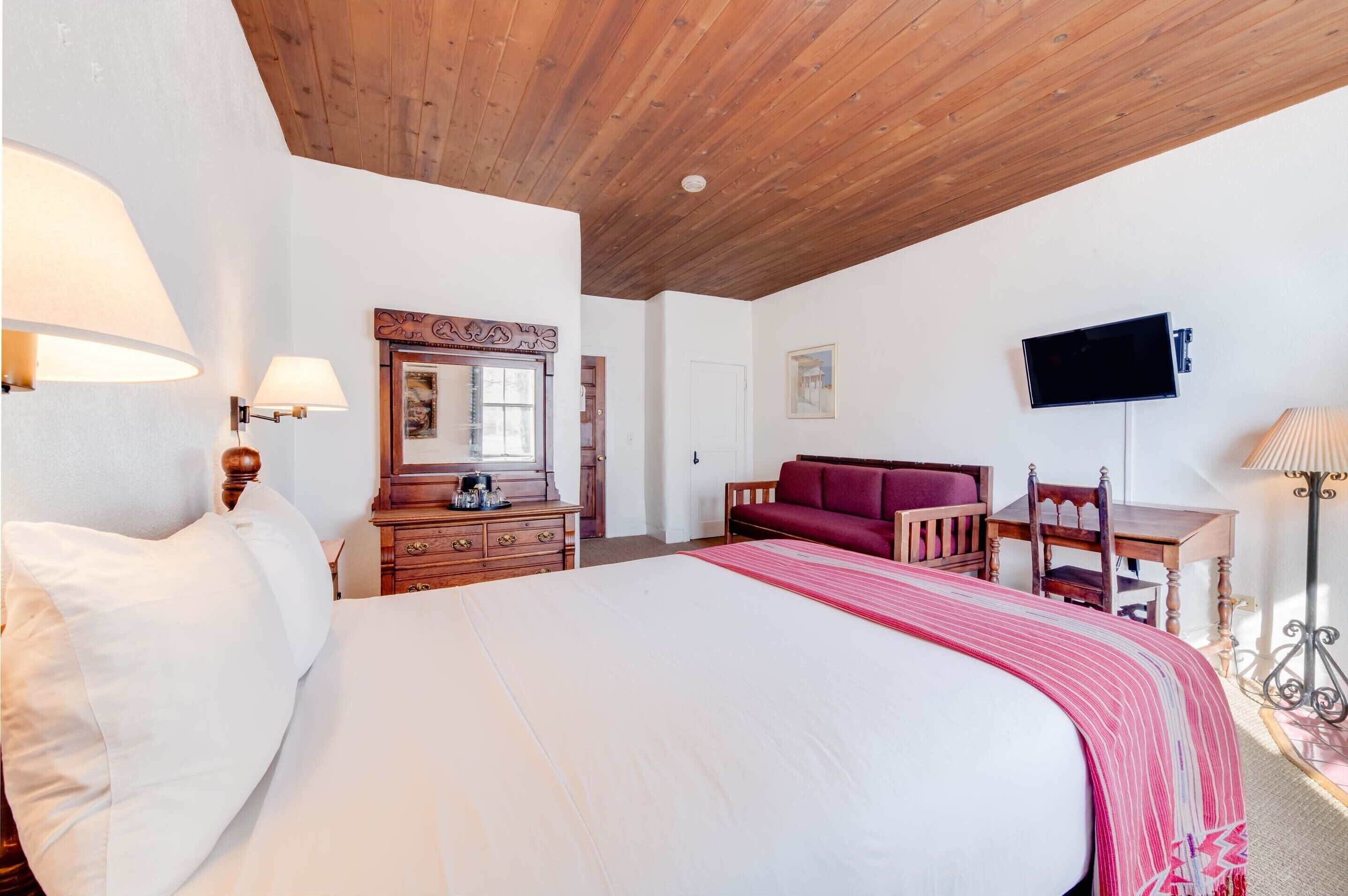 A bedroom with a large bed, wooden ceiling, a dresser, a red sofa, a TV mounted on the wall, and a desk with a chair.