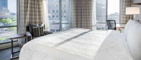Habitación de hotel con una cama grande, un escritorio, una silla y vistas a la ciudad a través de amplios ventanales.