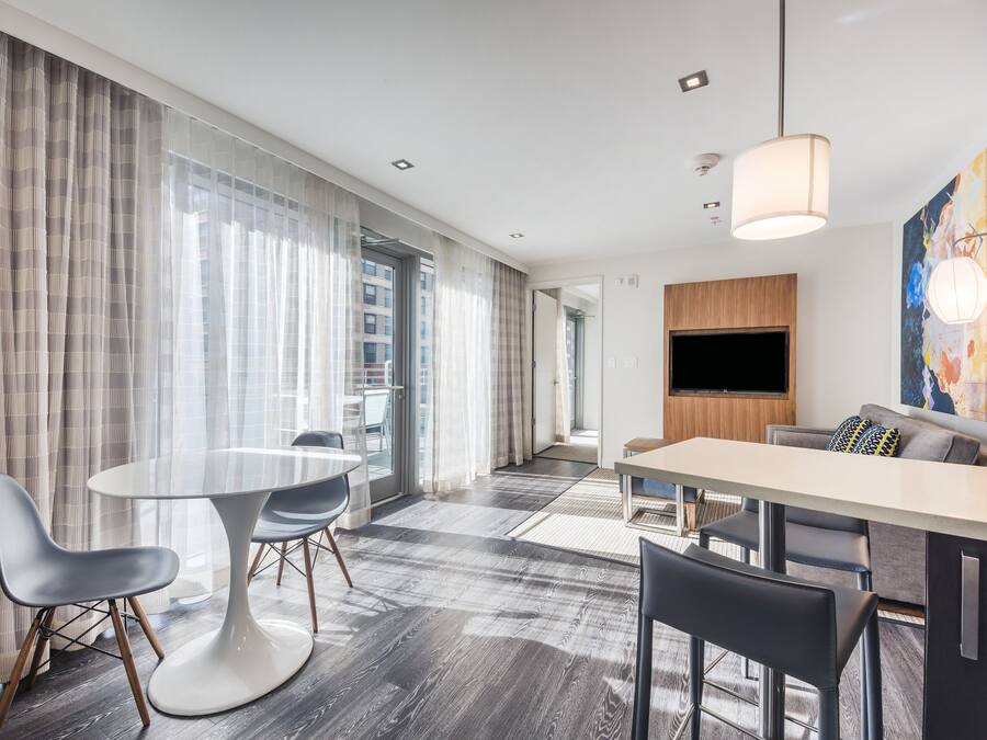 Een moderne hotelkamer met een zithoek, een bank, een aan de muur bevestigde televisie en grote ramen met gordijnen.