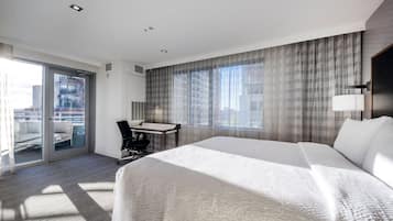 A hotel room with a large bed, a desk, a chair, and a view of the city through large windows.