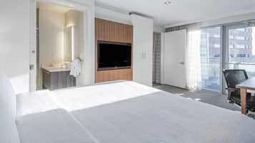 Una habitación de hotel moderna con una cama grande, un televisor montado en la pared, un baño con espejo y lavamanos, y un balcón con vistas a la ciudad.