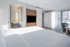 Una habitación de hotel moderna con una cama grande, un televisor montado en la pared, un baño con espejo y lavamanos, y un balcón con vistas a la ciudad.