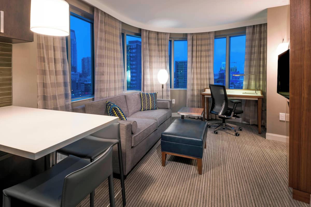 A modern hotel room with a curved layout, a sofa, a desk, a chair, and a TV. Large windows offer a city view.