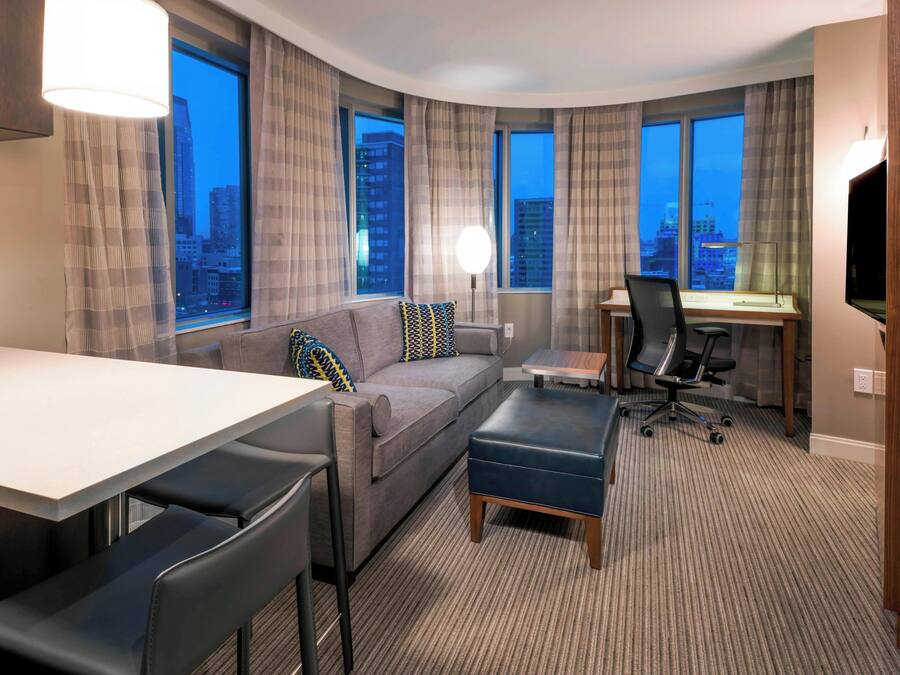A modern hotel room with a curved layout, a sofa, a desk, a chair, and a TV. Large windows offer a city view.