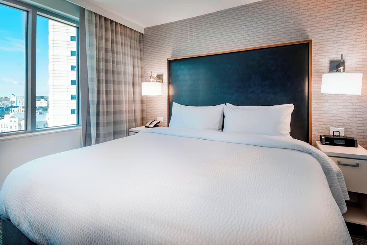 A hotel room with a large bed, a nightstand with a phone, a lamp, and a view of the city through a window.