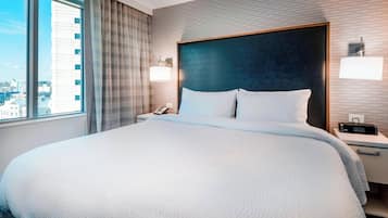Habitación de hotel con una cama grande, una mesita de noche con un teléfono, una lámpara y vistas de la ciudad a través de una ventana.