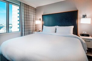 Habitación de hotel con una cama grande, una mesita de noche con un teléfono, una lámpara y vistas de la ciudad a través de una ventana.