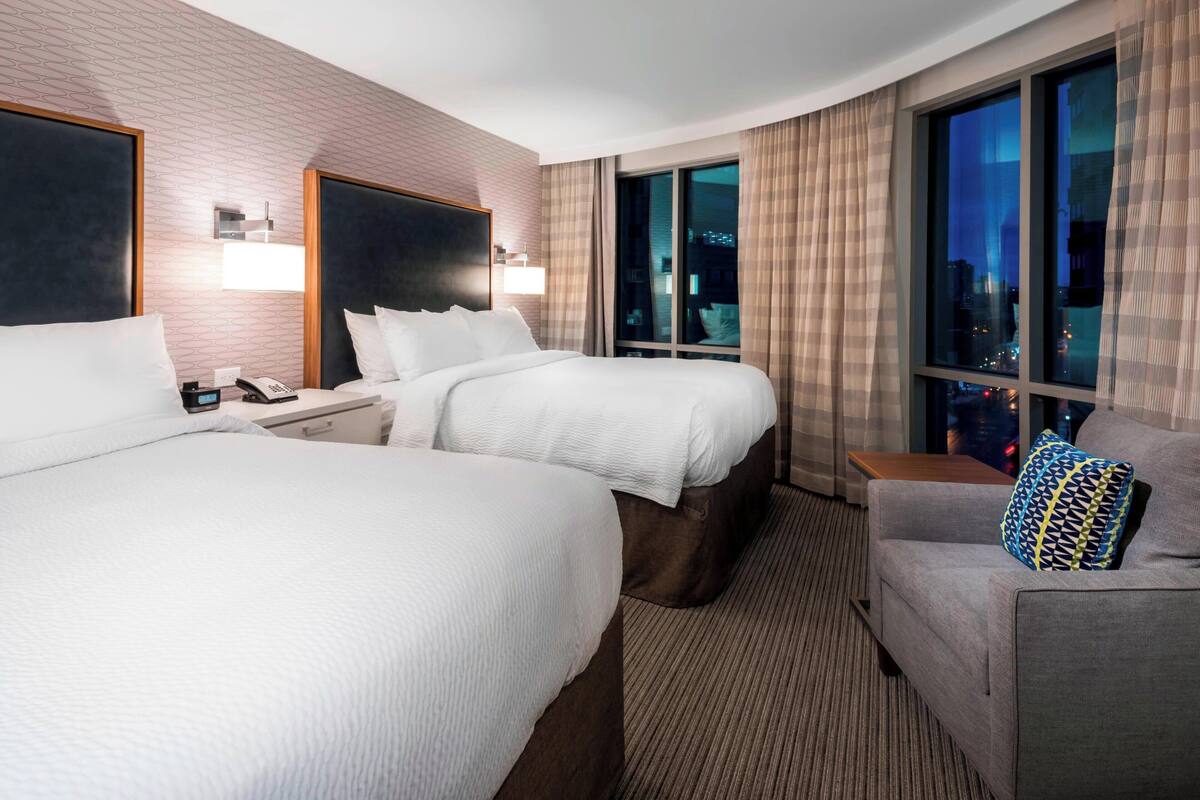A hotel room with two beds, a sofa, a nightstand, and a large window with a city view.