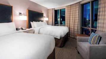 Ein Hotelzimmer mit zwei Betten, einem Schlafsofa, einem Nachttisch und einem großen Fenster mit Blick auf die Stadt.