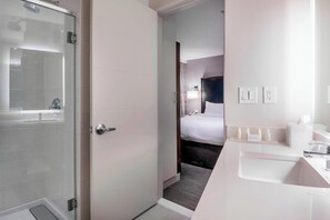 A modern hotel bathroom with a glass shower enclosure, a white sink, and a bed visible through an open door. - Residence Inn by Marriott Jersey City (Jersey City)