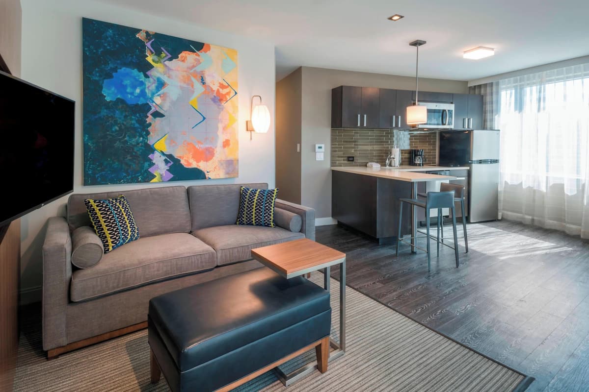 A modern living room with a sofa, ottoman, and a coffee table. There is a kitchen area with a microwave, refrigerator, and bar stools. A large abstract painting is on the wall, and a flat-screen TV is mounted on the wall.