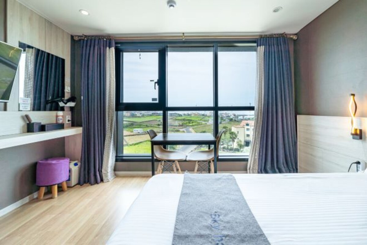 Una habitación de hotel con una cama grande, un escritorio con una silla, un taburete morado pequeño y un ventanal con vistas al campo.