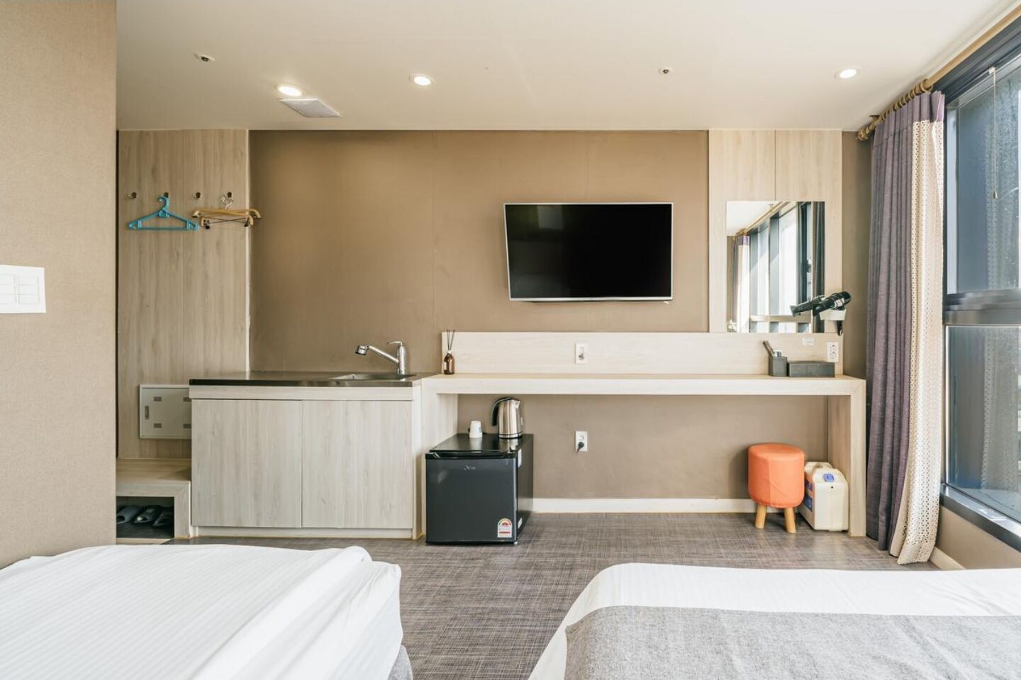 Ein Hotelzimmer mit zwei Betten, einer Küchenzeile, einem Flachbildfernseher, einem kleinen Hocker und einem Fenster mit Vorhängen.