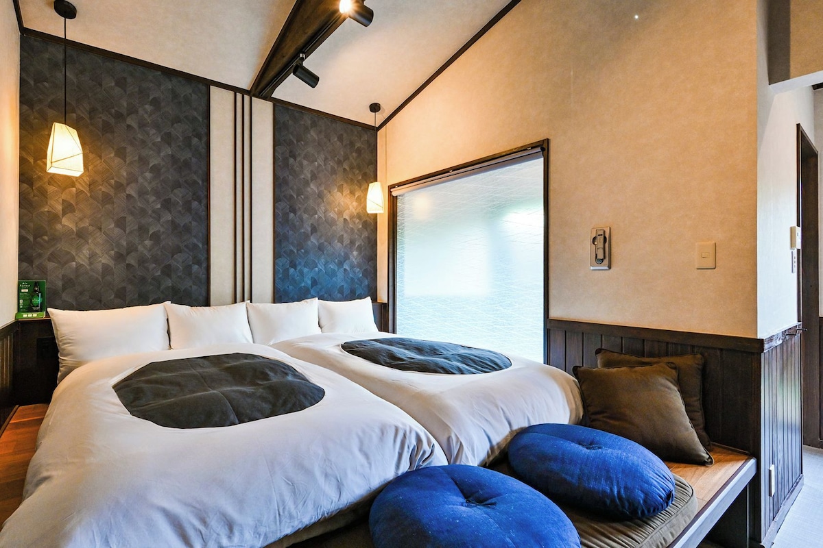 Quarto de hotel moderno com uma cama grande, duas mesas de cabeceira, uma janela com vista e um aparelho de luz fixado na parede.