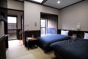 Ein Hotelzimmer mit zwei Betten, einem Schreibtisch, einem Sessel, einem kleinen Tisch, einem Fenster und einer Tür, die nach draußen führt.