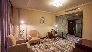 A hotel room with a carpeted floor, two armchairs, a small table with a plant, a desk with a chair, and a glass partition wall.