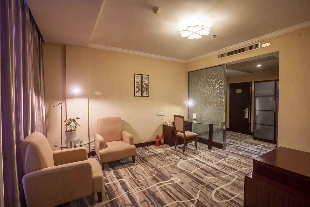 Ein Hotelzimmer mit Teppichboden, zwei Sesseln, einem kleinen Tisch mit Pflanze, einem Schreibtisch mit Stuhl und einer Glastrennwand.