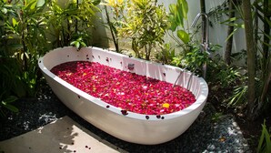 A white bathtub filled with rose petals, surrounded by lush greenery and a concrete wall. - Villa Senara (Canggu)