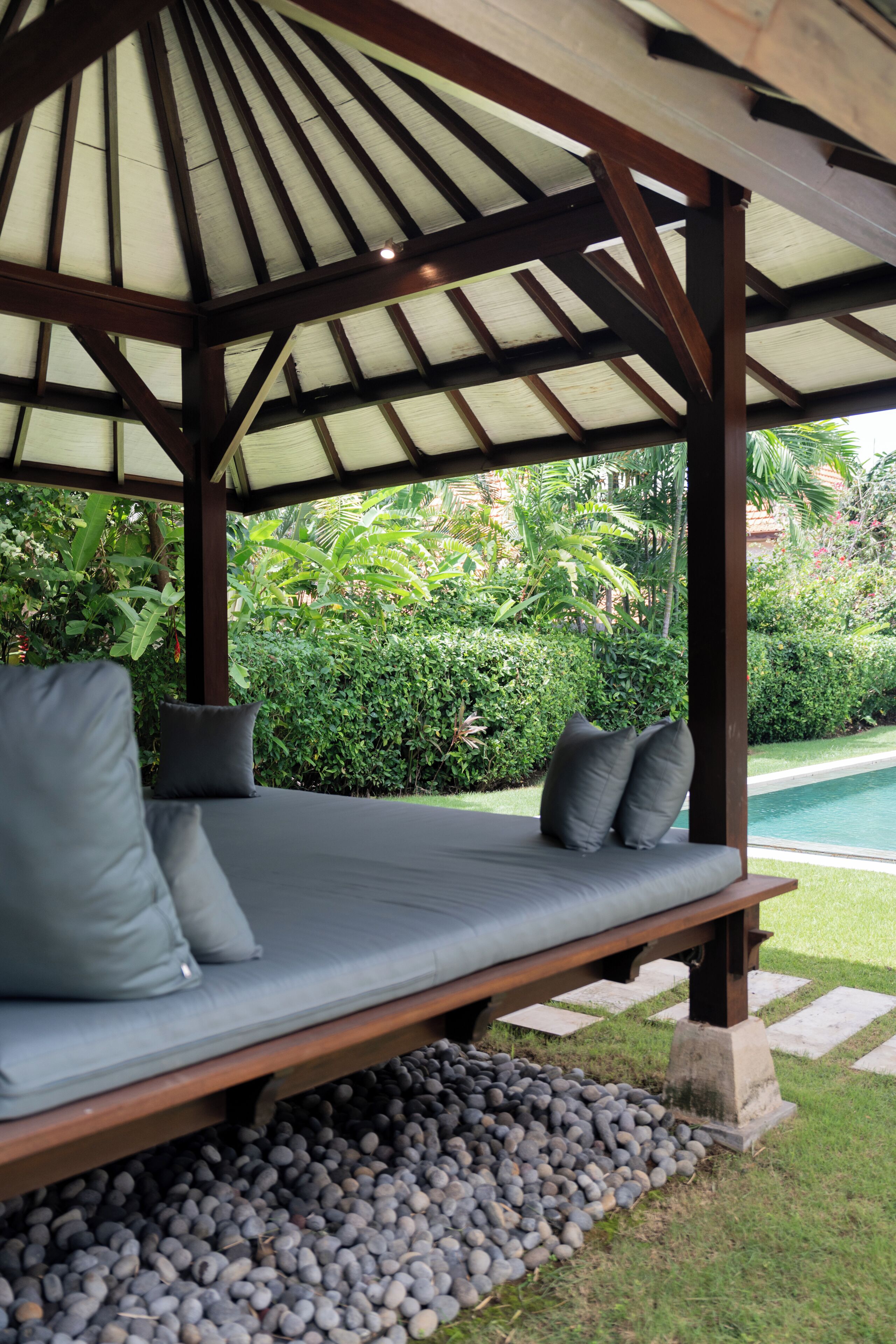 A covered outdoor seating area with a bench and cushions, surrounded by a garden and a pool.