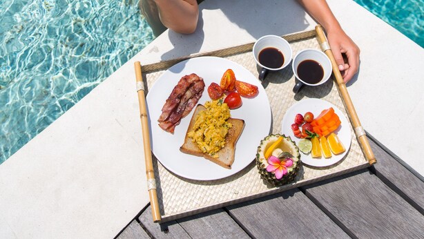 A person sitting by a pool with a tray of breakfast food including toast, bacon, eggs, tomatoes, and fruit. - Villa Senara (Canggu)