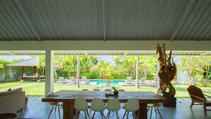 An outdoor dining area with a wooden table and chairs, a pool in the background, and a tropical garden view. - Villa Senara (Canggu)