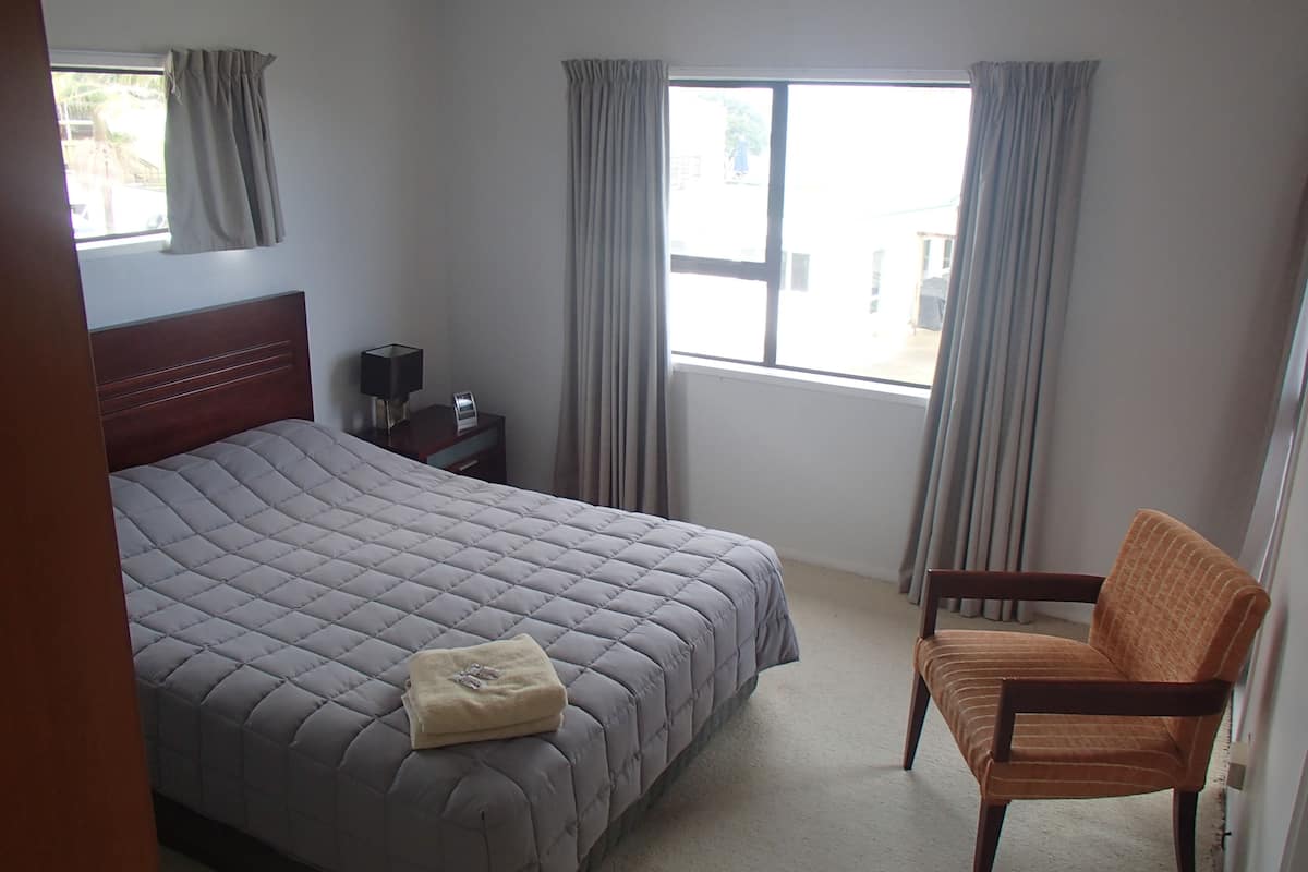 Habitación de hotel con una cama grande, una mesita de noche de madera, una silla y una ventana con cortinas.