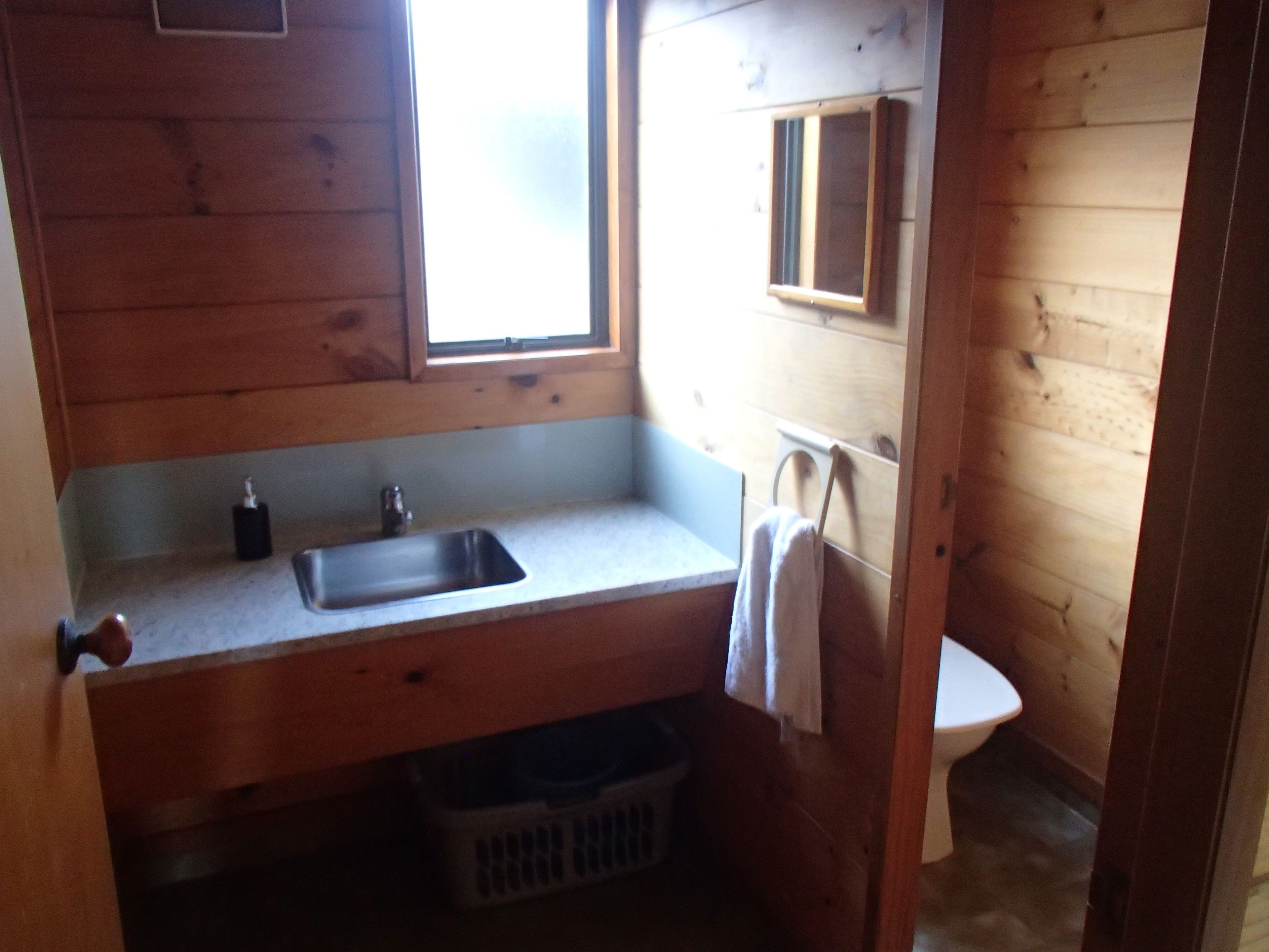 A small, wooden cabin-style bathroom with a single sink, a window, and a toilet.