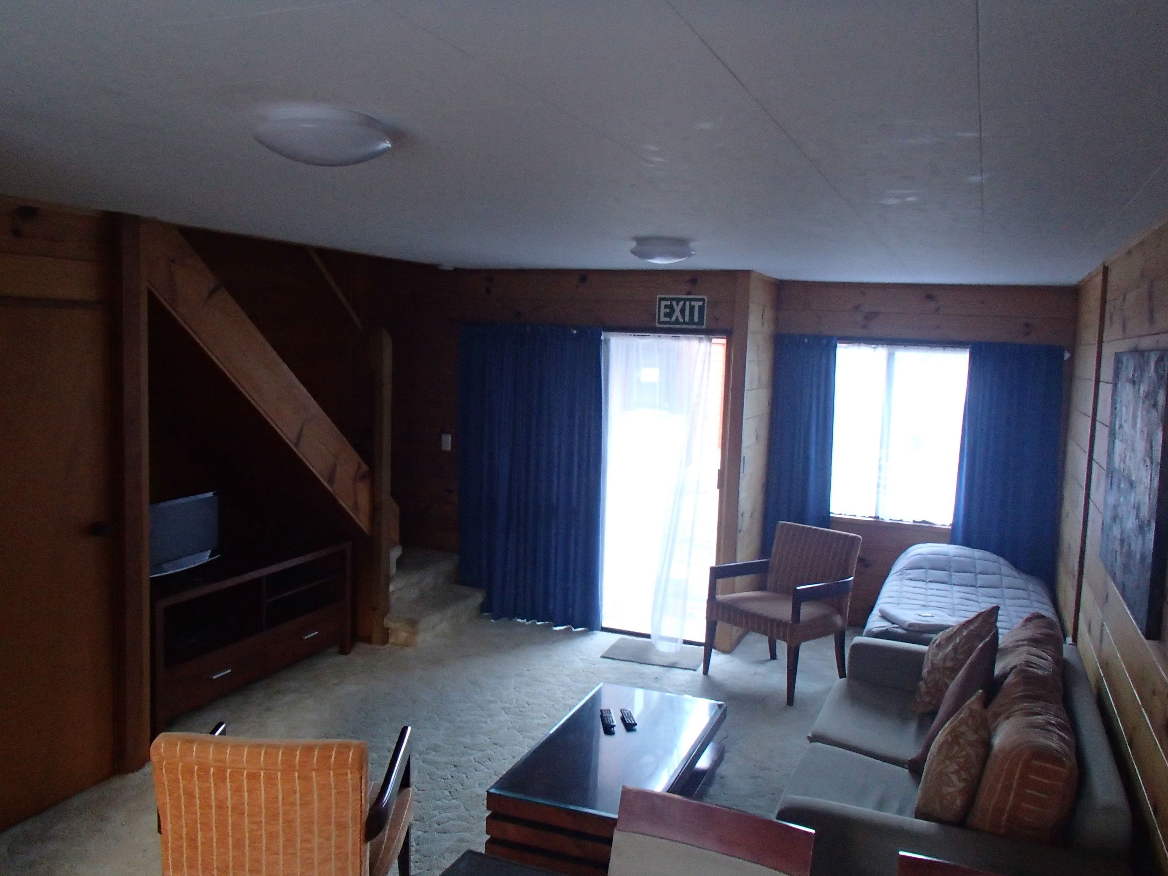 A cabin interior with a bed, a sofa, a TV, a table, and an exit sign.