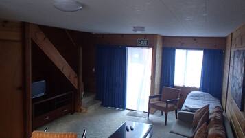 A cabin interior with a bed, a sofa, a TV, a table, and an exit sign.