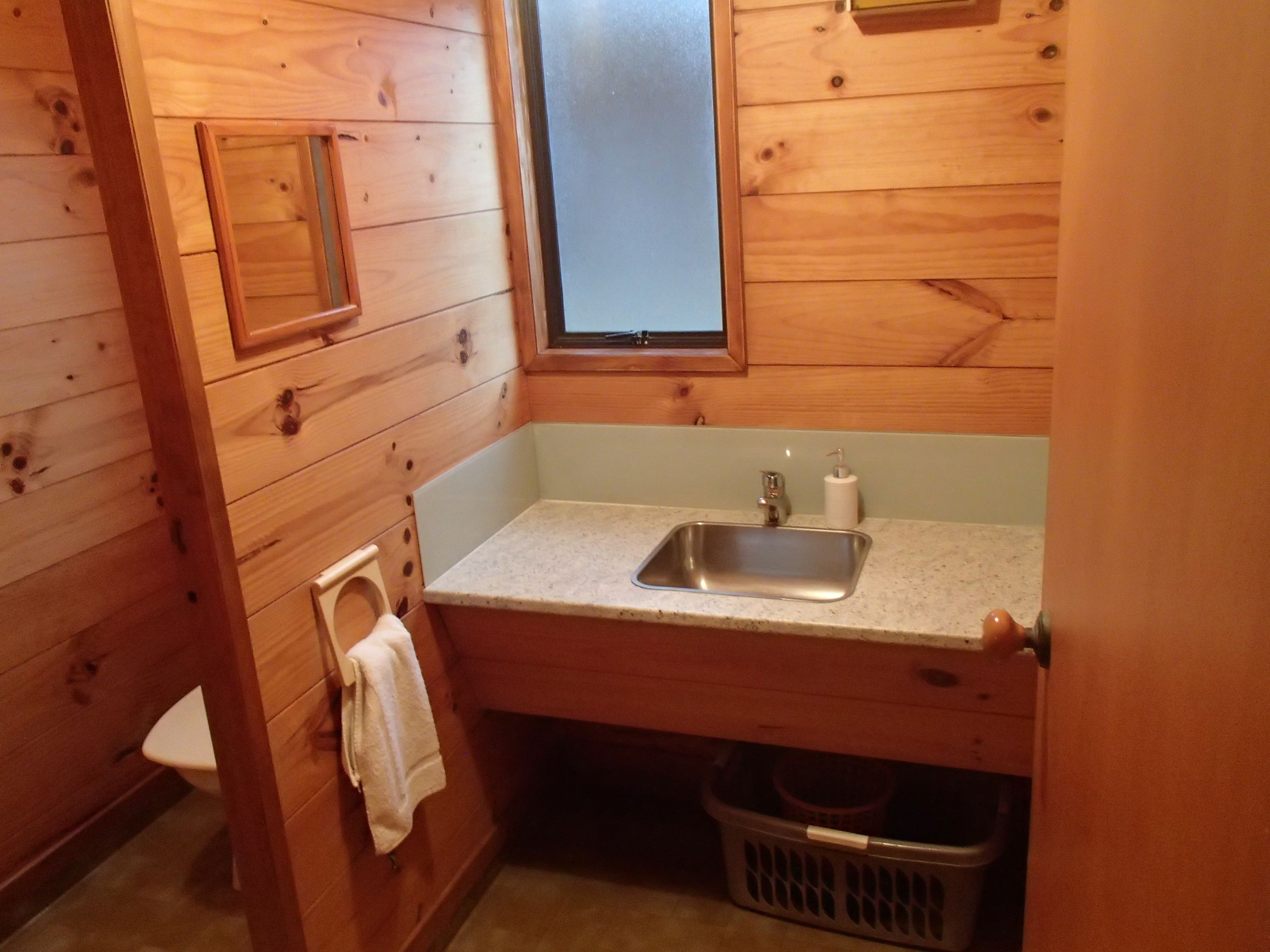 A small, wooden cabin-style bathroom with a single sink, a toilet, and a window above the sink.