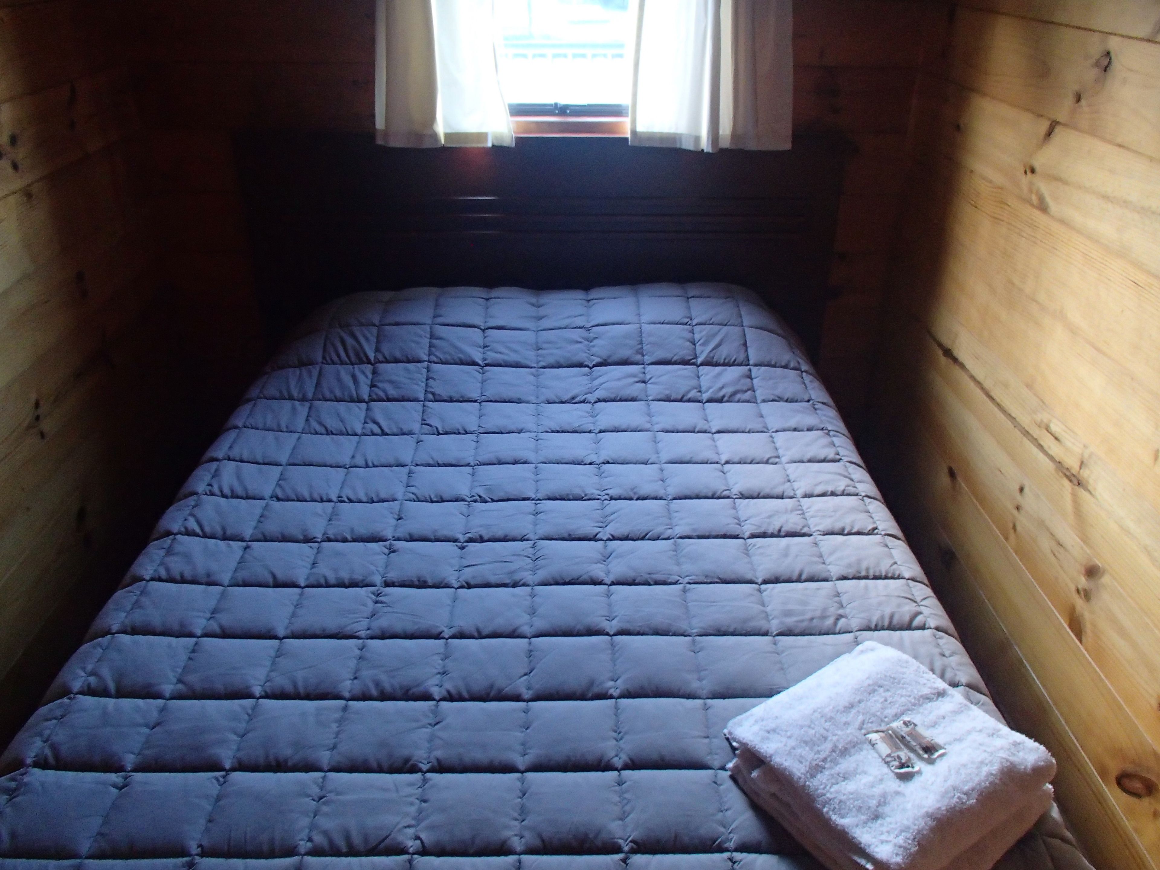 A narrow room with a single bed, wooden walls, and a window with curtains.