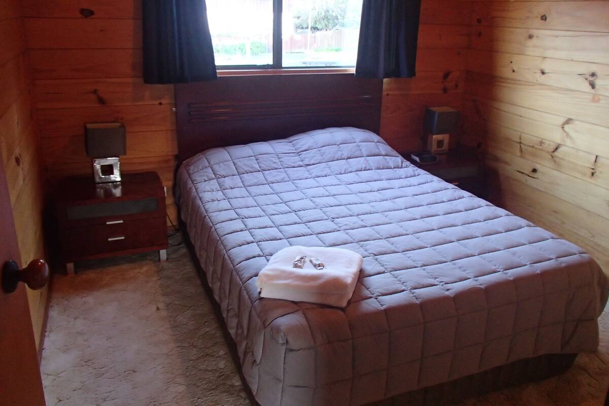 Habitación de cabaña de madera con cama, mesita de noche, lámpara y ventana con cortinas.