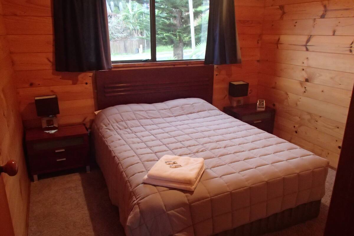 Un dormitorio de cabaña de madera con una cama, dos mesitas de noche y una ventana con cortinas.
