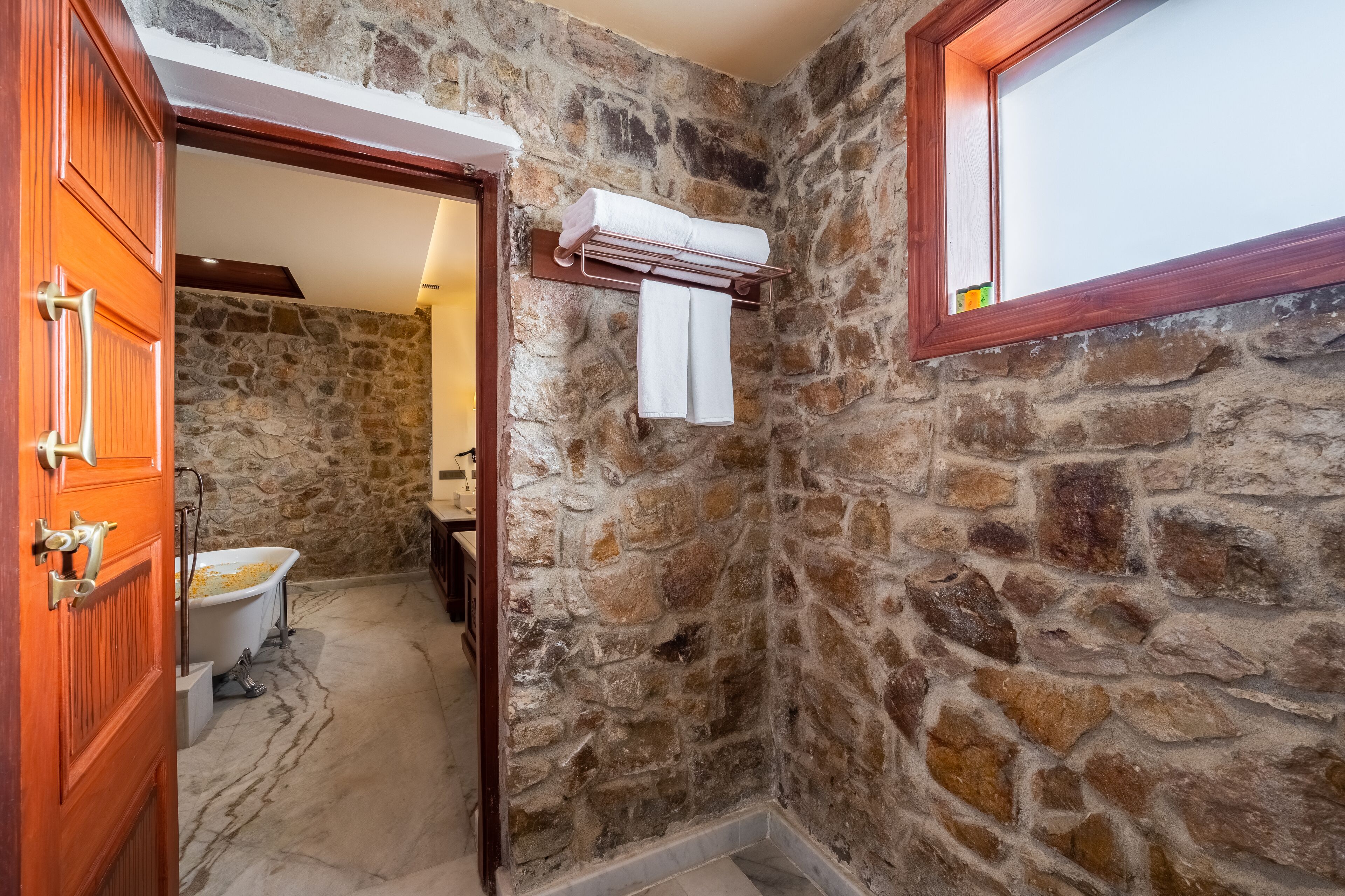 A stone-walled bathroom with a bathtub, a towel rack, and a wooden door.