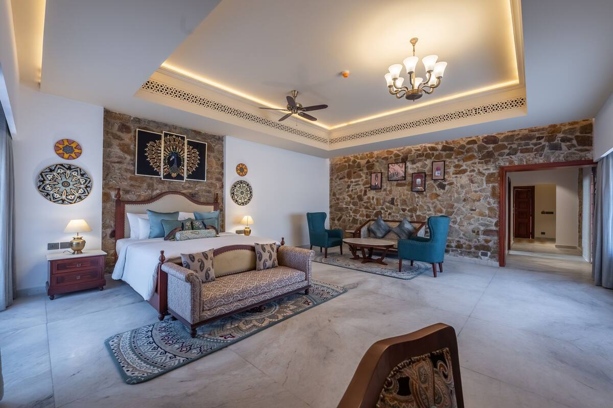 Ein geräumiges Schlafzimmer mit einem großen Bett, einer Sitzecke mit einer Couch und einem Esstisch. Das Zimmer verfügt über eine Steinwand, einen Deckenventilator und dekorative Wandbilder.