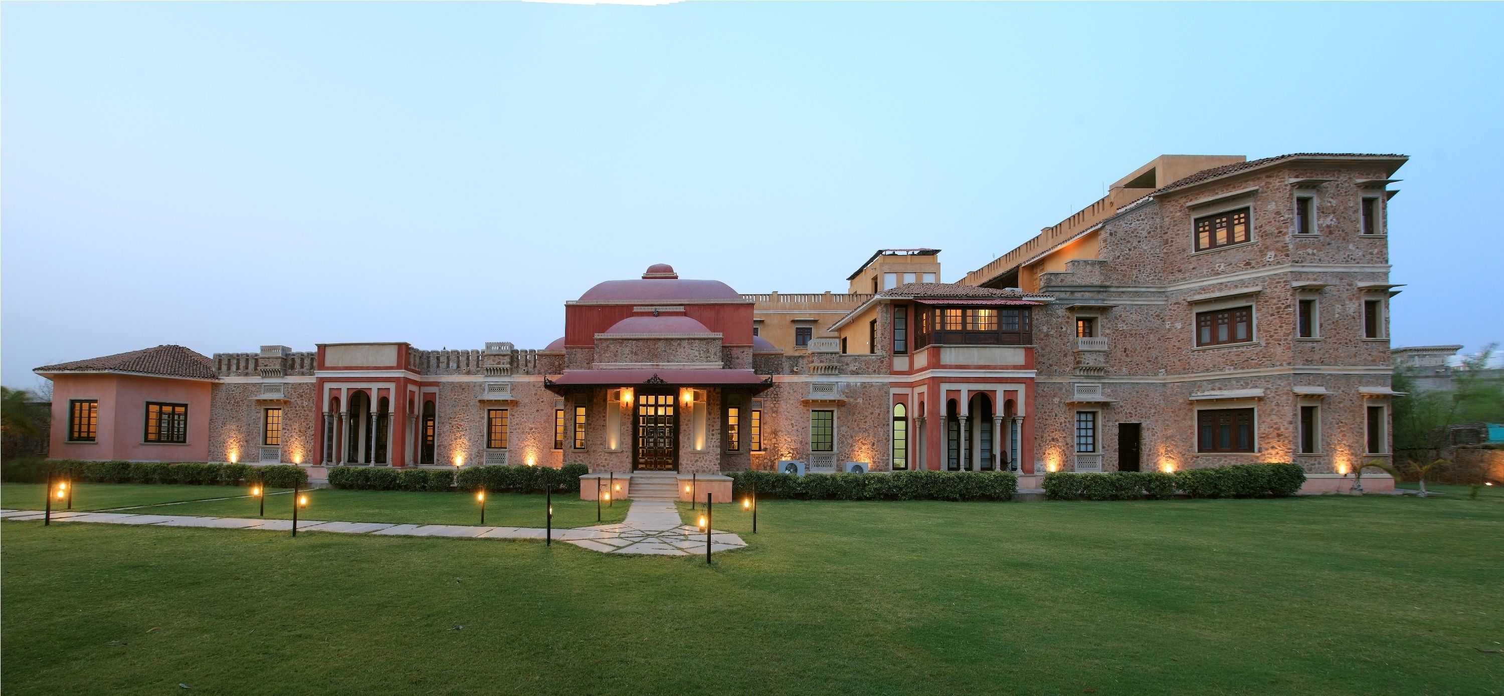 A large, multi-story building with a central dome and multiple wings, featuring a well-maintained lawn and outdoor lighting.
